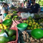 A man stands behind a table full of fresh fruits and vegetables, including bananas, oranges, and apples.