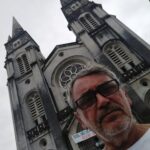 A man stands in front of a large church, wearing glasses and a white shirt.