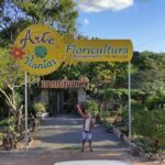 A man stands in front of a sign for Arte Floricultura, holding a purple object.