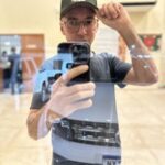A man wearing a green hat and glasses takes a picture of his reflection in a mirror.