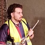 A man wearing a yellow vest is holding a pipe.