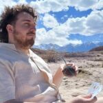 A man with a beard and long hair is sitting in the desert, holding a cup and a deck of cards.