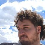 A man with curly hair and a beard is looking up at the sky, possibly admiring the clouds.