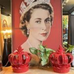 A painting of Queen Elizabeth II hangs on a wall, surrounded by two red vases.