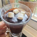 A person holds a cup of chocolate with marshmallows on top, ready to enjoy a delicious treat.