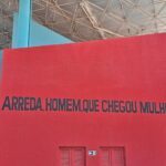 A red wall with a black sign that says "Arreda, homo, que chego de mulher".