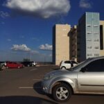A silver car is parked in a parking lot, with a large building in the background.