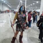 A woman dressed in a Scottish outfit, complete with a blue face paint, holds a sword and stands in a large room.
