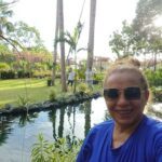 A woman in a blue shirt is smiling and taking a selfie in front of a pond.