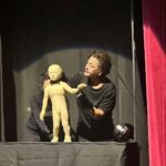 A woman is holding a doll in front of a curtain, possibly preparing for a performance or a presentation.