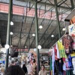 A woman stands in a market, surrounded by various items for sale.