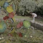 A young girl stands near a pond, surrounded by colorful umbrellas and flowers.