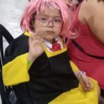 A young girl wearing a pink wig and glasses is sitting in a chair.