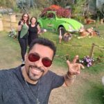 Three men pose in front of a green Volkswagen Beetle, which is parked on a grassy area.
