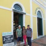 Three people pose in front of a yellow building, with one of them wearing a hat.