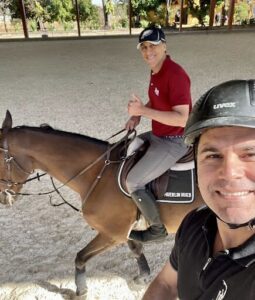 Two men are riding horses, one wearing a helmet and the other giving a thumbs up.