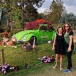 Two women pose in front of a green Volkswagen Beetle, which is parked in a garden setting.