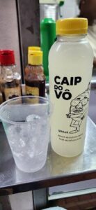A bottle of Caipirinha sits next to a glass of ice and a bottle of hot sauce.