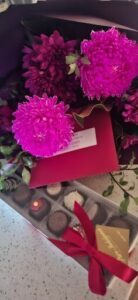 A bouquet of purple flowers sits in a red cardboard box, accompanied by a card.