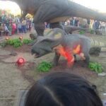 A group of people are gathered around a fake dinosaur exhibit, with a small dinosaur statue on display.