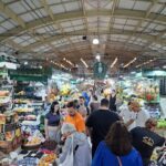 A group of people are shopping in a market, with a variety of fruits and vegetables on display.