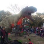 A large dinosaur statue is lit up at night, drawing a crowd of people who are standing around it.