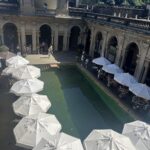A large group of people are gathered around a swimming pool, with many umbrellas providing shade.