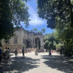 A large group of people are gathered in front of a building, possibly a castle, with trees in the background.