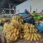 A large pile of bananas is displayed at a market, with a man standing nearby.