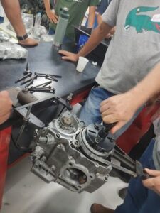 A man is working on a motorcycle engine, surrounded by other people who are watching the process.