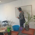 A man is writing on a white board while standing in front of a potted plant.