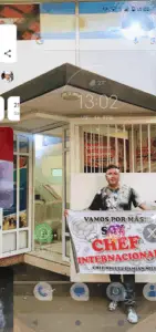 A man stands in front of a building holding a sign that says "Vamos por mas!".