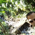A man wearing sunglasses and a green shirt stands in front of a waterfall.