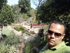 A man wearing sunglasses stands in front of a beautiful garden.