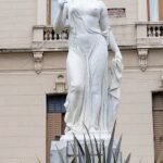 A statue of a woman holding a mirror is located in front of a building.
