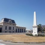 A white obelisk stands in front of a large building, possibly a museum or a government building.