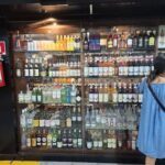 A woman is standing in front of a refrigerator filled with various bottles of alcohol.