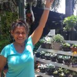 A woman is standing in front of a table full of potted plants, holding her arm up in the air.