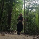A woman wearing a black dress stands in a forest, looking up at the sky.