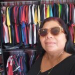 A woman wearing sunglasses stands in front of a clothing rack, showcasing her style and the variety of shirts available.