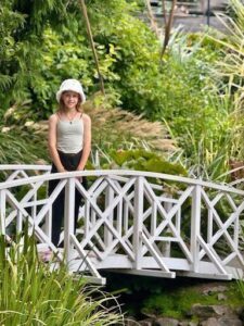 A young woman wearing a white hat stands on a bridge, surrounded by lush greenery.