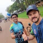 Two men wearing bicycle helmets and jerseys are standing on a road, one giving a thumbs up.
